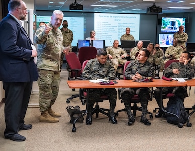 Soldiers sit in a classroom setting with screens in the background
