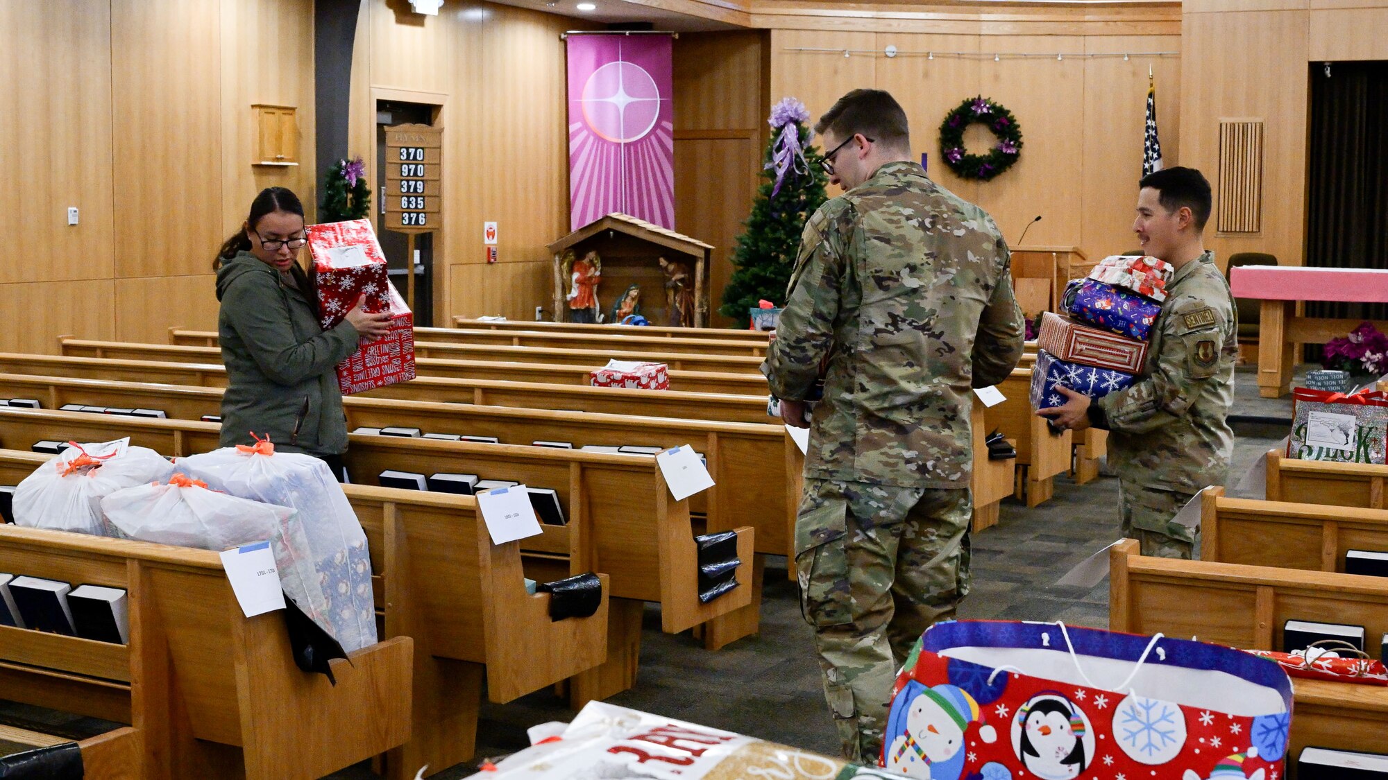 A woman in civilian attire and two men in uniform walk down pew aisles placing wrapped gifts.