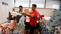 Two men in a room filled with wrapped gifts load presents into a shopping cart.