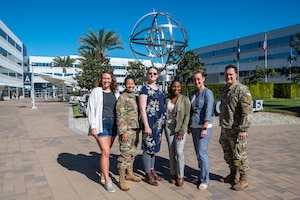 Five women, one in uniform, and one man in uniform stand in front of a sculpture in the courtyard of Space Systems Command.