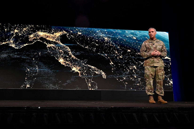 A commander stands on a stage in front of a screen displaying Europe at night from space.