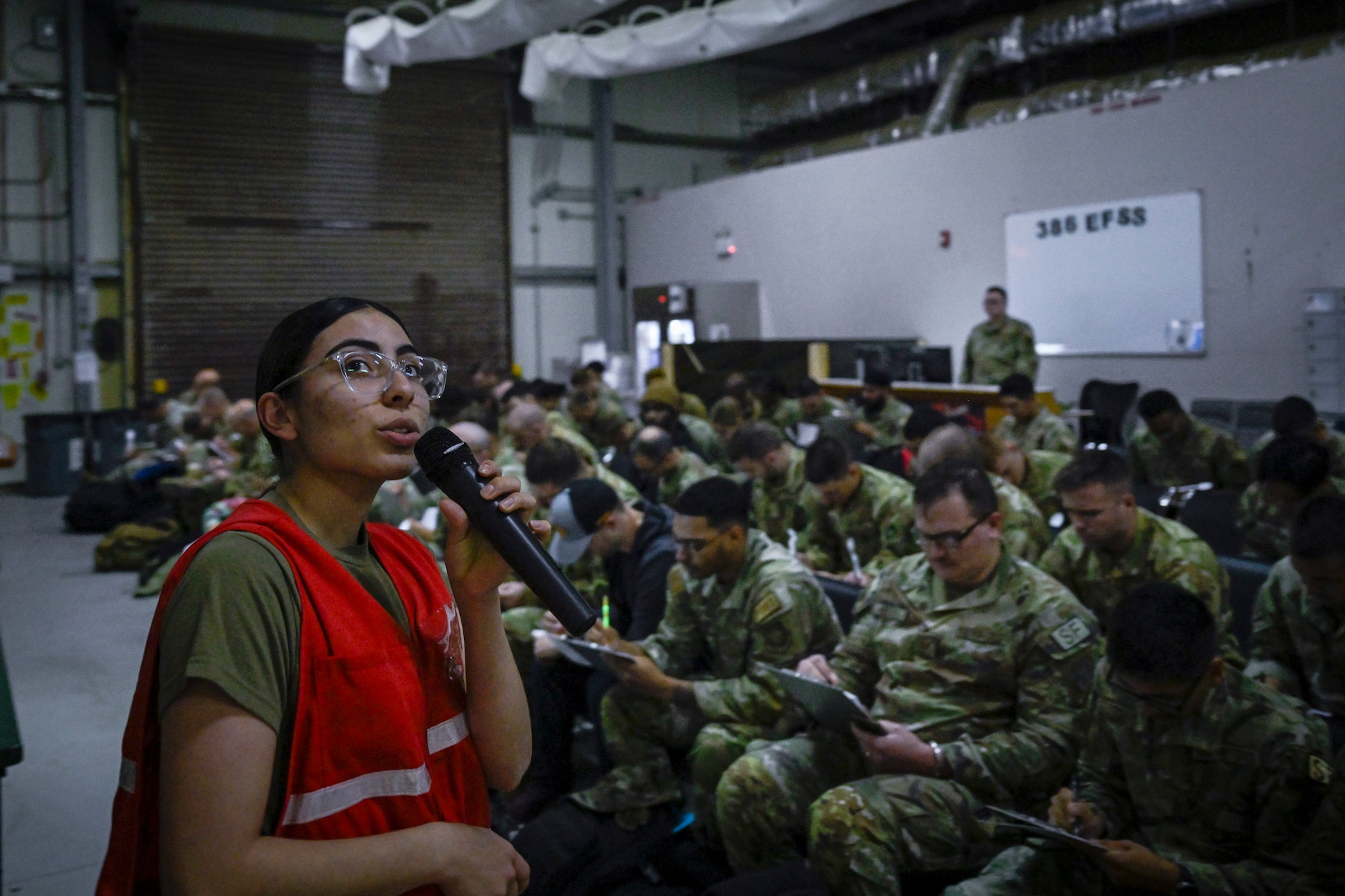 A military member wearing a red vest briefs newly arrived military personnel.