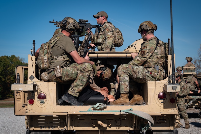 Service members sit in a military vehicle; one stands atop it manning a weapon.