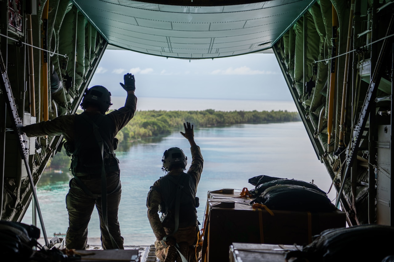 Two airmen wave from the back of an open aircraft loaded with supply boxes to an island below.