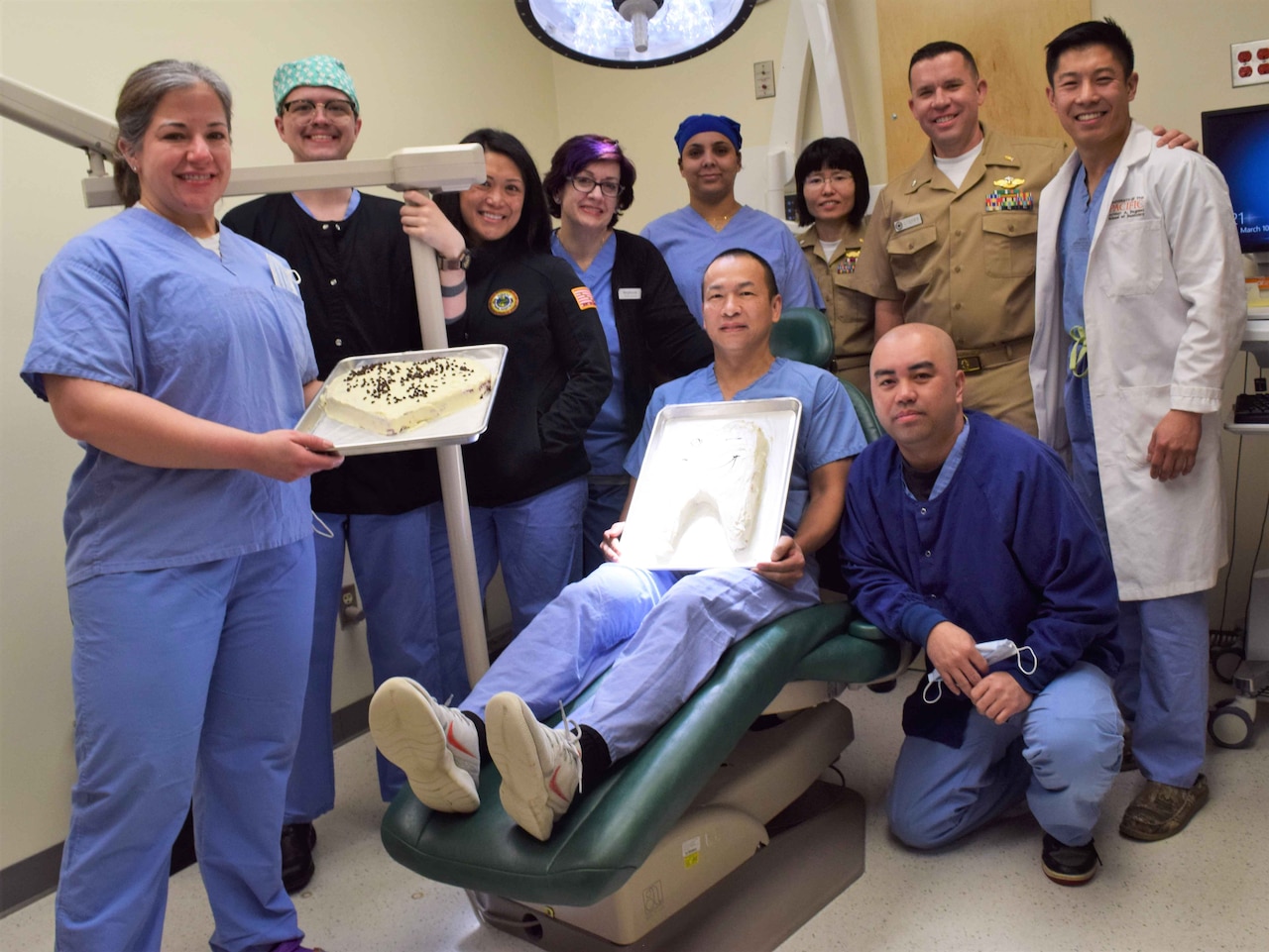 A group of people holding two cakes poses around a dentist’s chair.