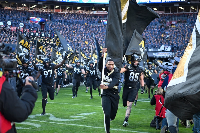 A cadet carrying an Army flag runs in front of football athletes in a stadium.