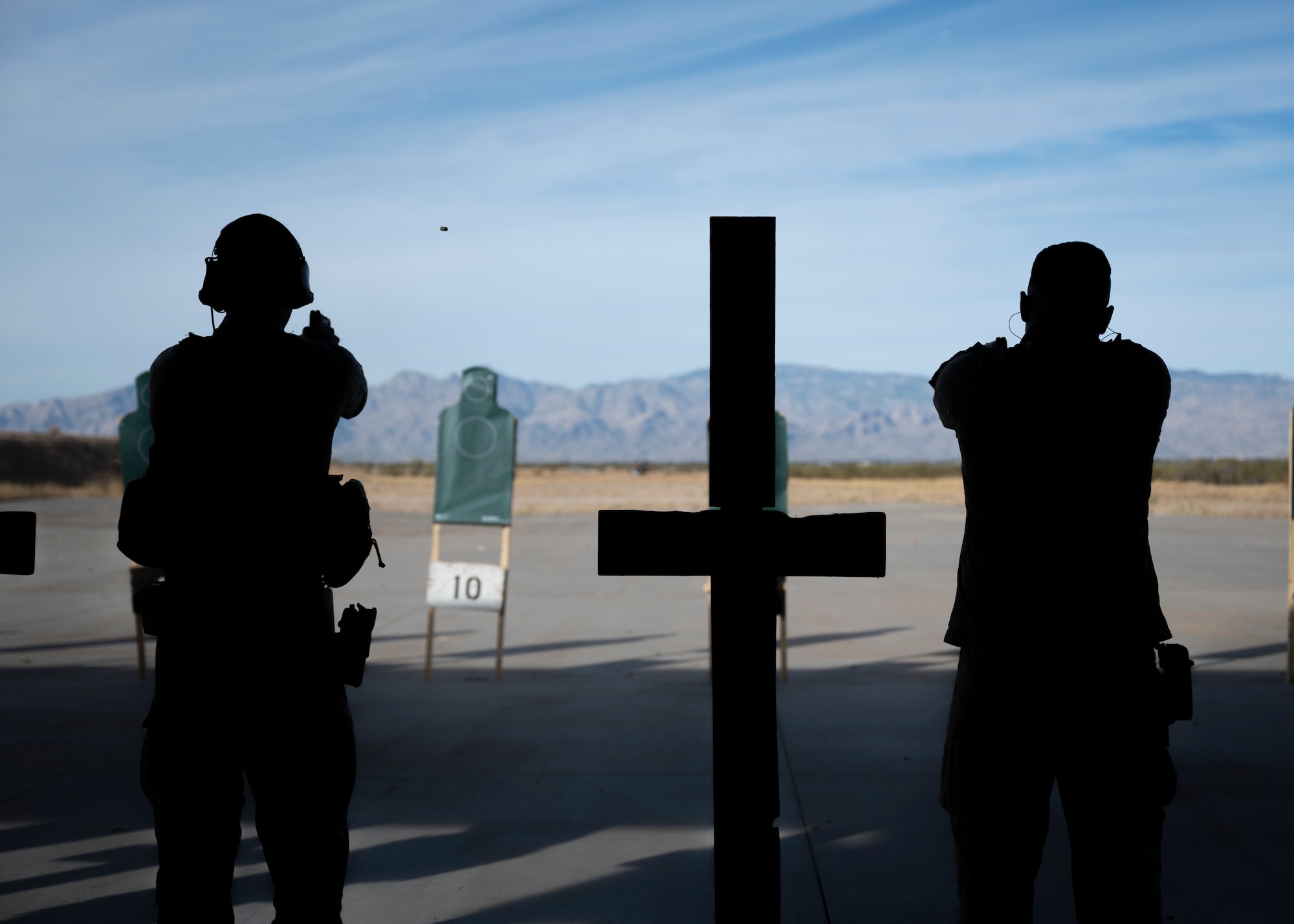 The silhouettes of two men in military uniforms aiming handguns at targets at a firing range.