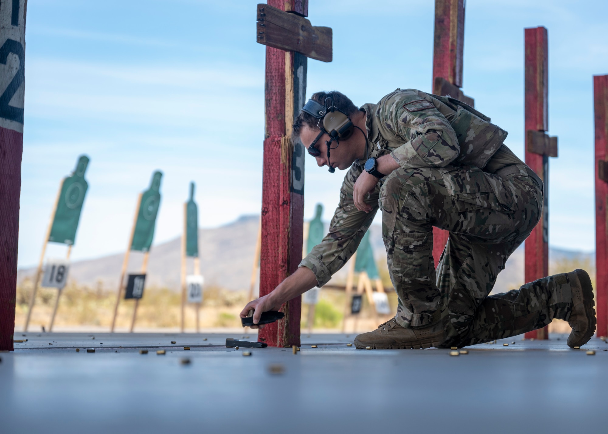 A man in military uniform kneels down to pick up used brass on a firing range.