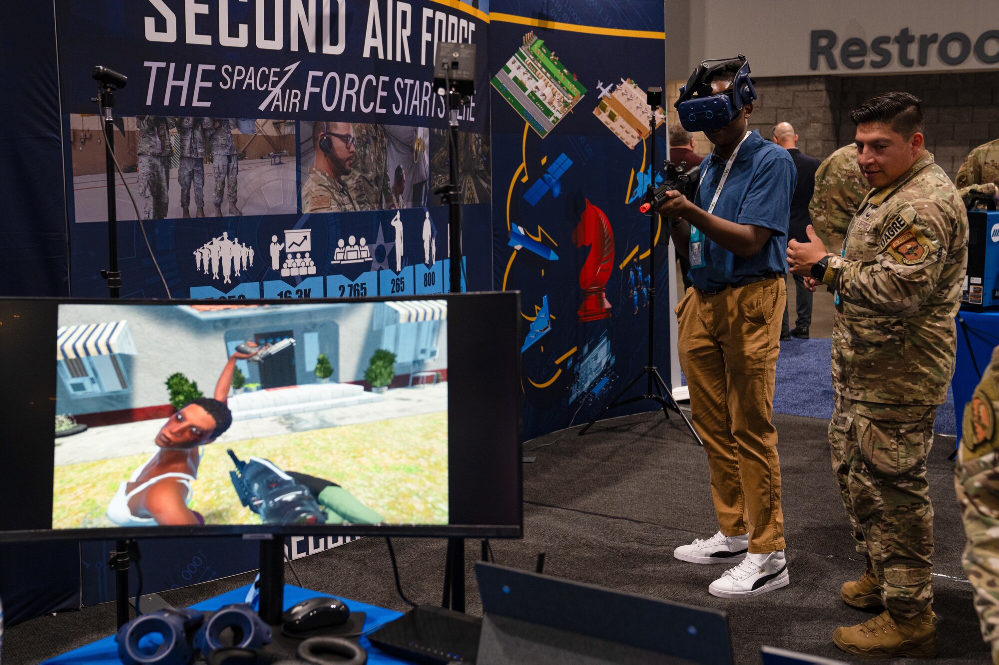 A conference attendee wears virtual reality goggle and holds a model assault rifle while being coach a man in current U.S. Air Force uniform.
