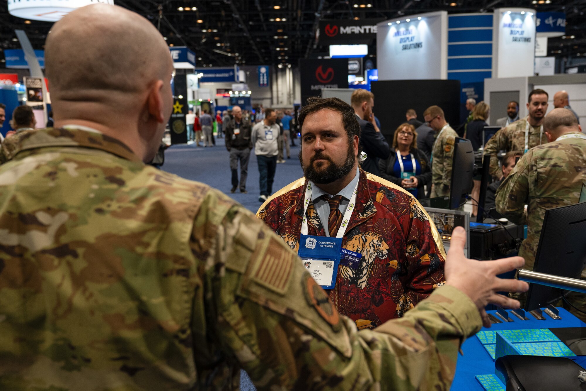 A man in operational camouflage pattern explains his booth to a conference attendee.
