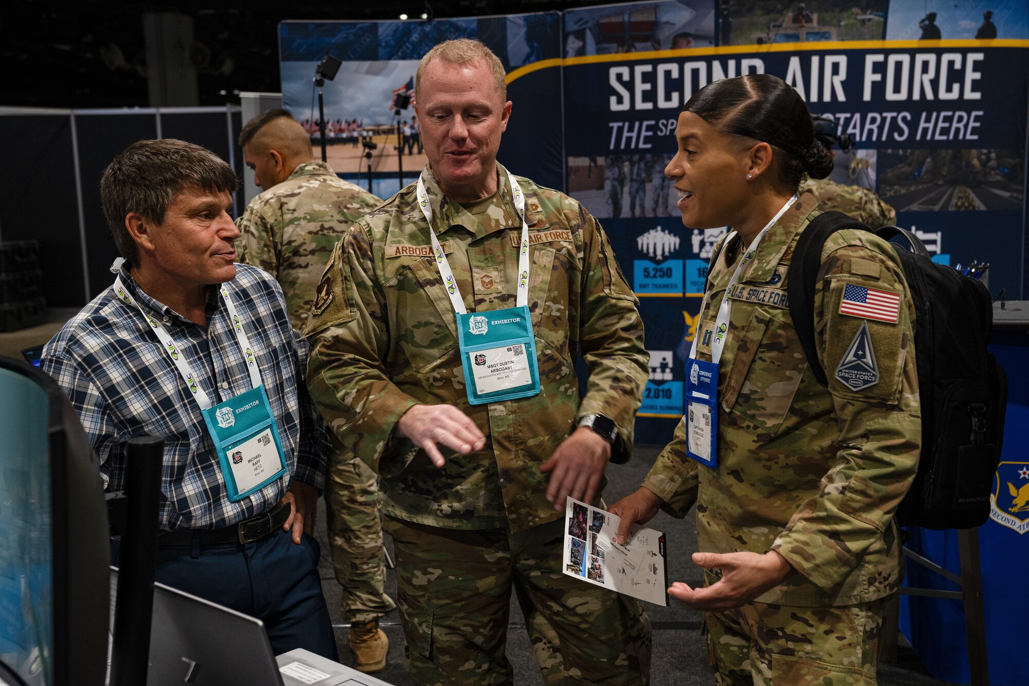 Shorter tan man on the left, tall white man in the middle and tan woman on the right have a conversation at booth at a conference.