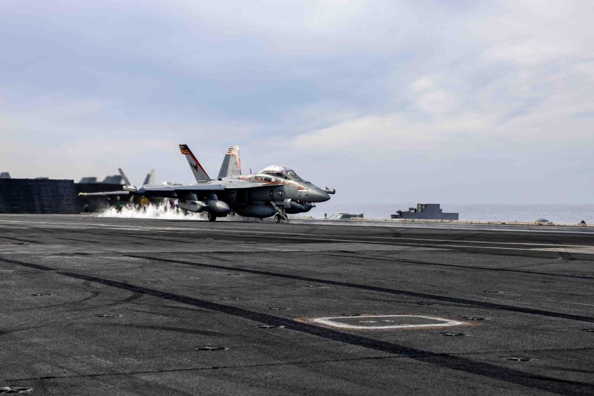 A fighter jet is taking off on a large ship flight line at sea. The ocean is in the background