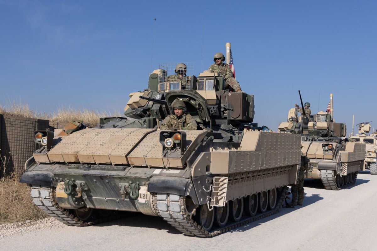 Men wearing camouflage uniforms and tactical helmets ride in and on a trio of military tanks lifting up American flags driving down a dirt road.