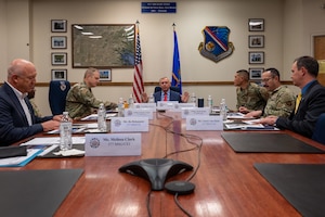 Men in suits and military members sit around at able with flags in the background