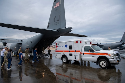 Military members walk towards the back of a C-130J Super Hercules and an ambulance.