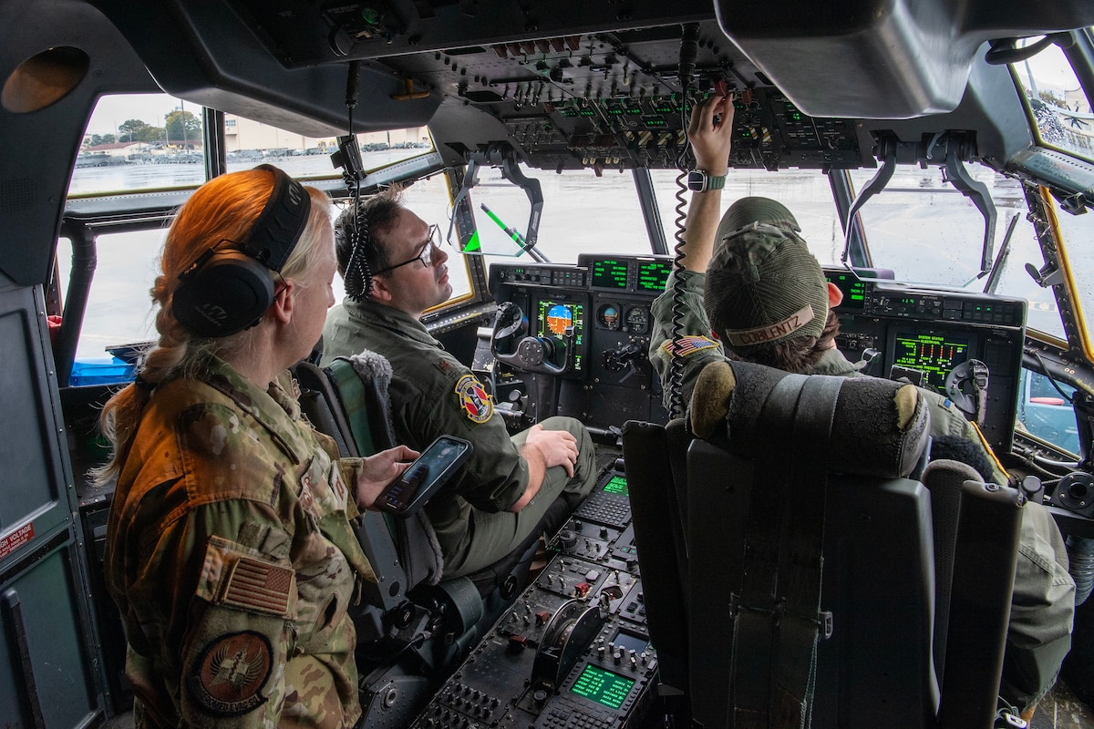 Three military members conduct preflight preparations in the cockpit of a C-130J Super Hercules.