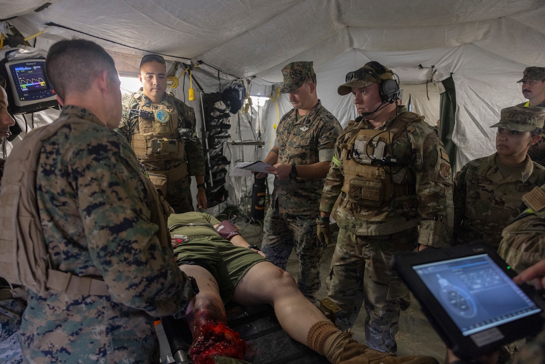 Military members assess simulated casualty on a stretcher inside a medical tent.