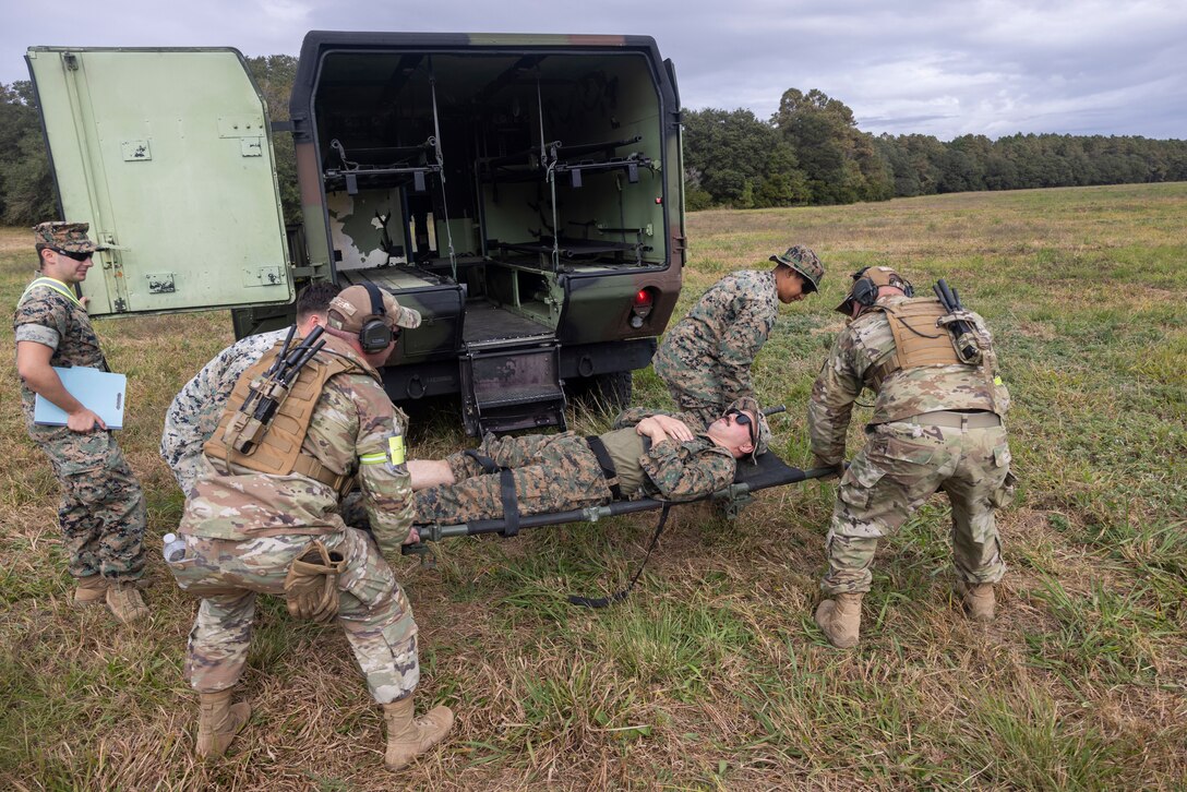 Four military members lift a simulated casualty on a stretcher to load into the back of a medical transport vehicle. One military member evaluating.