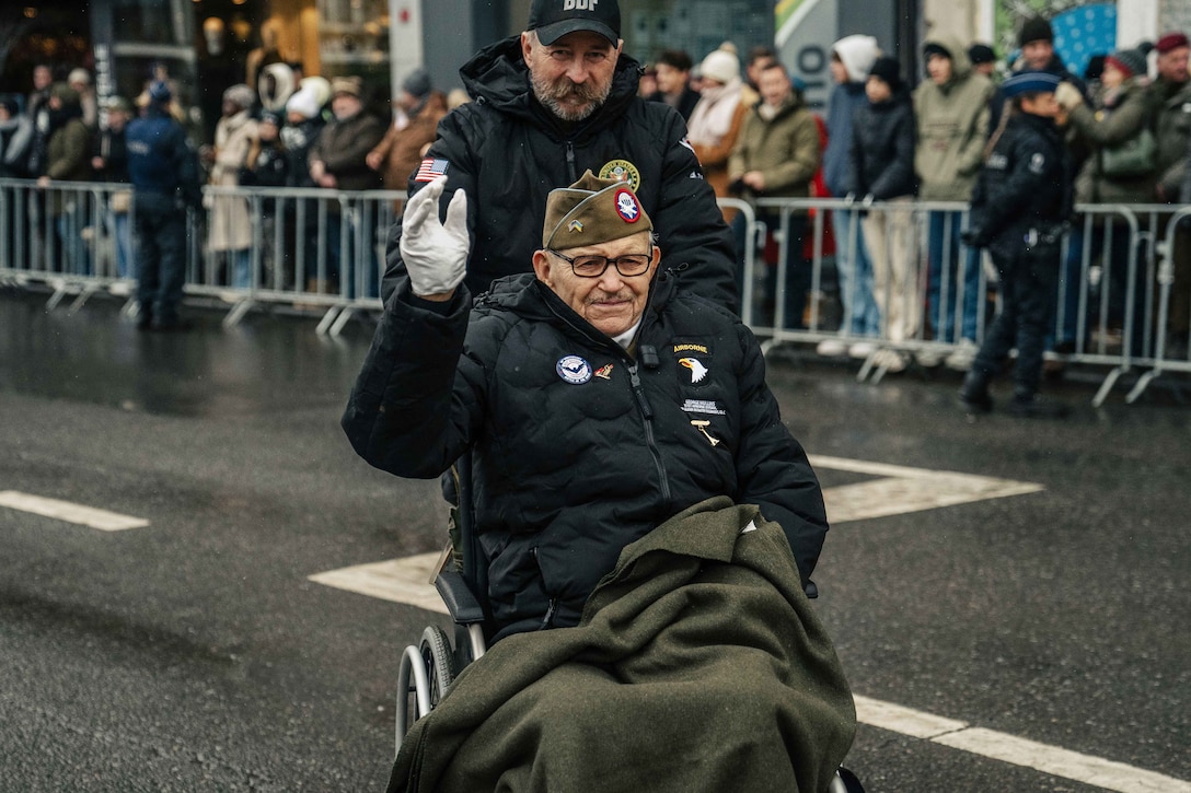 A veteran wearing a jacket and white gloves waves to the crowd while being pushed in a wheelchair along a street during a parade on a rainy day.