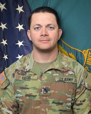 Man in U.S. Army uniform standing in front of two flags.