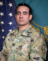 Man in U.S. Army uniform standing in front of two flags.