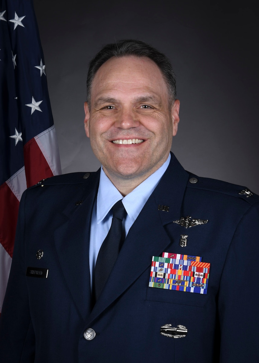 A portrait of a man in Air Force service dress with an American flag draped in the background.