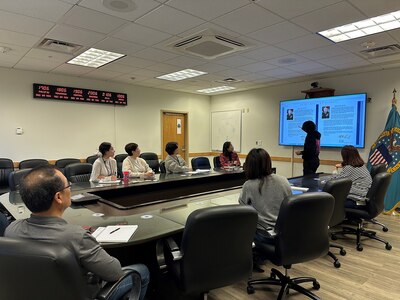 A group of people sit around a large conference table looking at a person who is pointing to a large video monitor on a wall.
