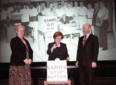 Two women and one man pose in front of a sign that says SAMMS "Stop" Button