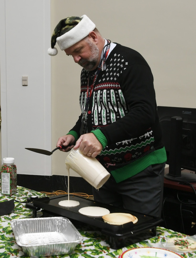 A man pours batter on to a grill. he is searing a Christmas sweater and Santa hat.