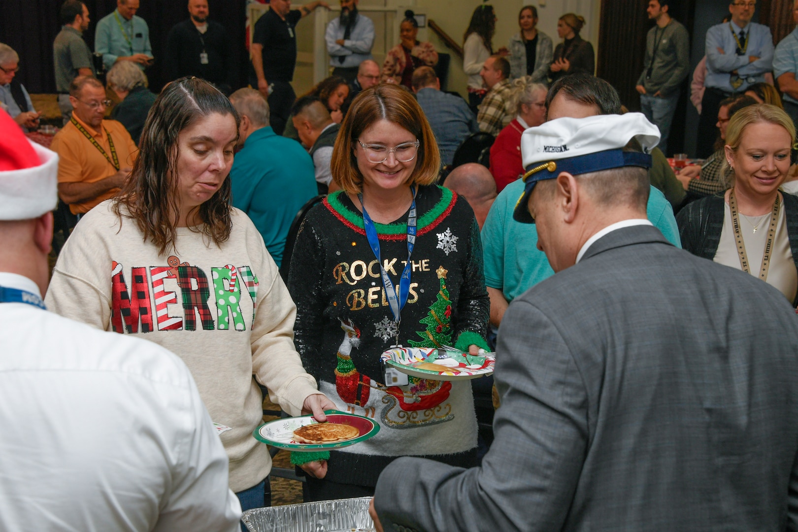 A man with his back to the camera serves food to two women wearing holiday sweaters.