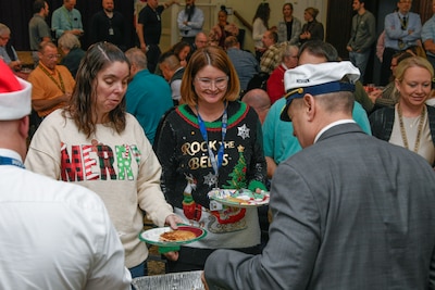 A man with his back to the camera serves food to two women wearing holiday sweaters.