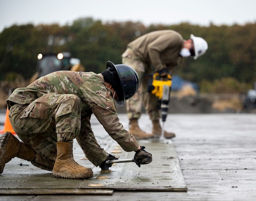 Two military members use equipment to drill and screw a piece of matting into the ground.