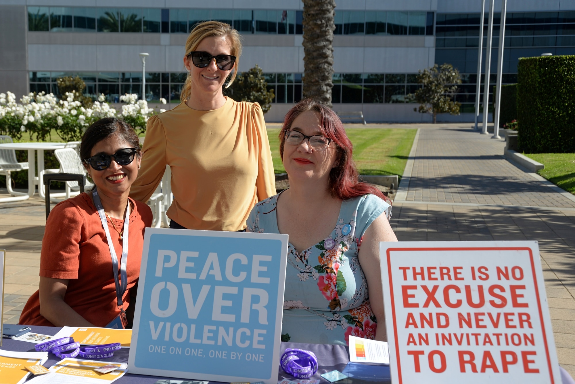 One woman stands while two women sit at a booth with two signs. One that reads, "Peace over violence," and the other says, "There is no excuse and never an invitation to rape."