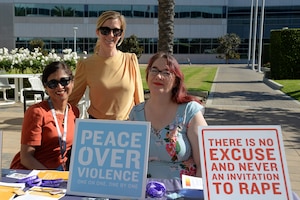 One woman stands while two women sit at a booth with two signs. One that reads, "Peace over violence," and the other says, "There is no excuse and never an invitation to rape."