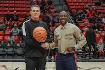 U.S. Marine Corps Sgt. Dennis Maina, assistant for Officer Selection Team San Diego, Recruiting Station San Diego,  poses for a photograph with a referee during the San Diego State University Men's Basketball game in San Diego, California, Dec. 11, 2024. Maina was recognized as the guest of honor during the game. The guest of honor received special recognition including a formal introduction, reserved seating and participation in game-day activities. (U.S. Marine Corps photo by Cpl. Alejandro Fernandez)