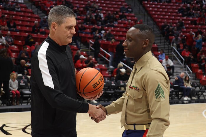 U.S. Marine Corps Sgt. Dennis Maina, assistant for Officer Selection Team San Diego, Recruiting Station San Diego,  presents the game ball to the referee during the San Diego State University Men's Basketball game in San Diego, California, Dec. 11, 2024. Maina was recognized as the guest of honor during the game. The guest of honor received special recognition including a formal introduction, reserved seating and participation in game-day activities. (U.S. Marine Corps photo by Cpl. Alejandro Fernandez)