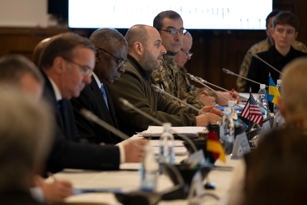 Man wearing business attire and foreign military uniforms are seated at a table with microphones.