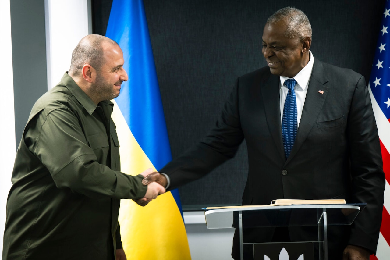 Two men shake hands while standing in front of U.S. and Ukrainian flags.