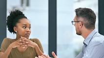 A young black woman and a middle-aged white male sitting at a desk having a conversation with papers sitting in front of them