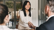 A young Asian female wearing a white blouse and slacks is leaning over a desk to shake hands with two NSA hiring managers