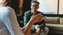 A woman with her back towards the camera having a conversation with a middle-aged white man who is sitting on a couch during an NSA interview