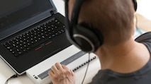 A man sitting at a desk with a computer, using a braille reader to understand what is on the screen while completing his NSA application