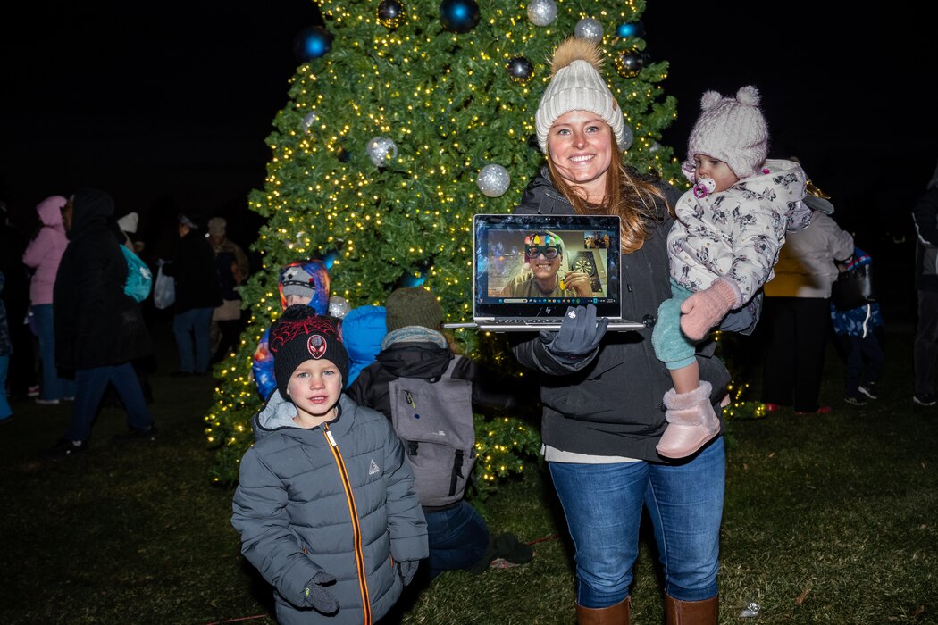 A woman, in winter coat and hat, holds her baby daughter, bundled in winter gear, while also holding a laptop with Capt. Dennis video calling on the screen, beside their son, in winter coat and cap