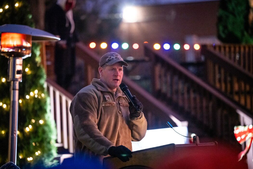 Col. Richards, in military fleece jacket and black gloves, holds a microphone behind a podium in front of lit Christmas decorations