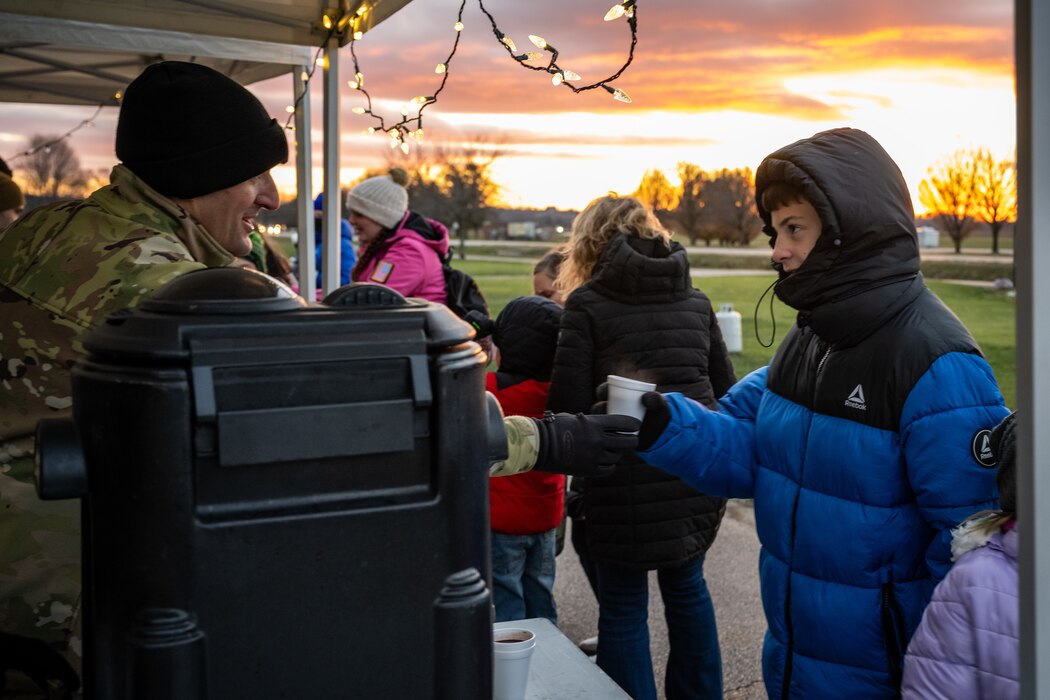 Chaplain Breznau, in military uniform and black winter cap, hands a white styrofoam cup to a young man, in blue winter coat and hood