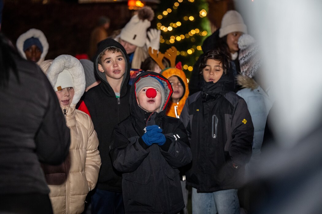 A group of young children wearing winter coats, one with a foam red nose and another with a set of fake antlers, sing in front of lit Christmas decorations