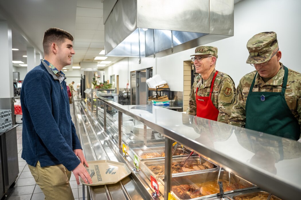 Gen. Richardson, in military uniform and Santa hat, smiles while looking over a metal meal prep counter, as Chief Master Sgt. Fitch, in military uniform and Santa hat, looks down prepping a meal for a young man, in blue jacket and khaki pants