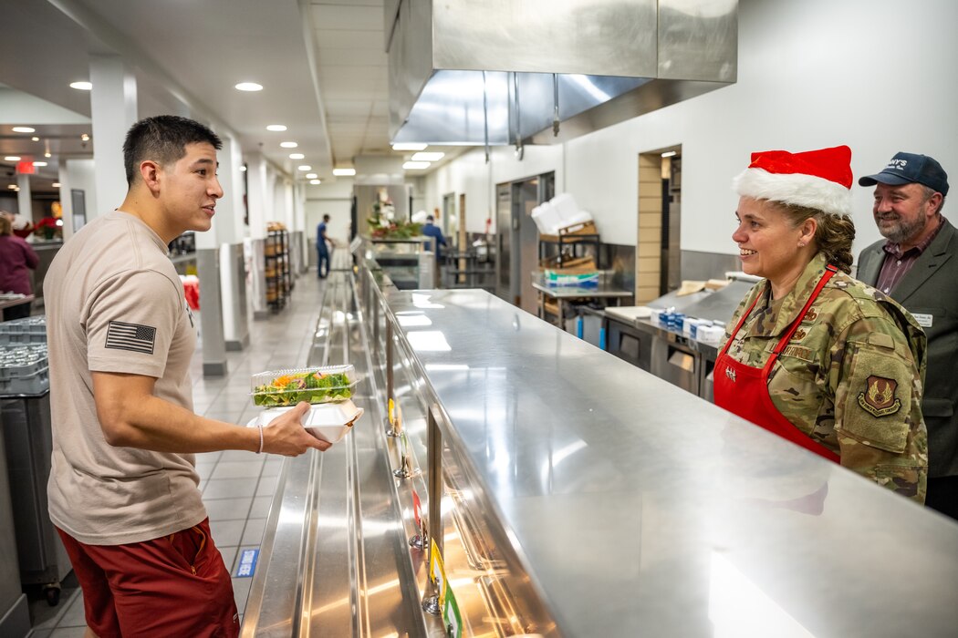 Lt. Gen. Hurry, in military uniform and Santa hat, smiles while looking over a metal meal prep counter at a young man, in tan tshirt with black American flag logo on sleeve