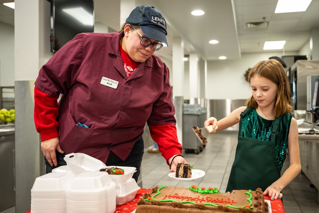 A young girl, in green dress and green apron, handles a serving spatula as a female employee, in maroon uniform and black cap, holds a plate with a chocolate piece of cake