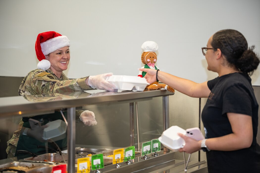 Chief Master Sgt. Fontaine, in military uniform and Santa hat, hands a styrofoam tray over a metal meal prep counter to a young woman, in black polo shirt